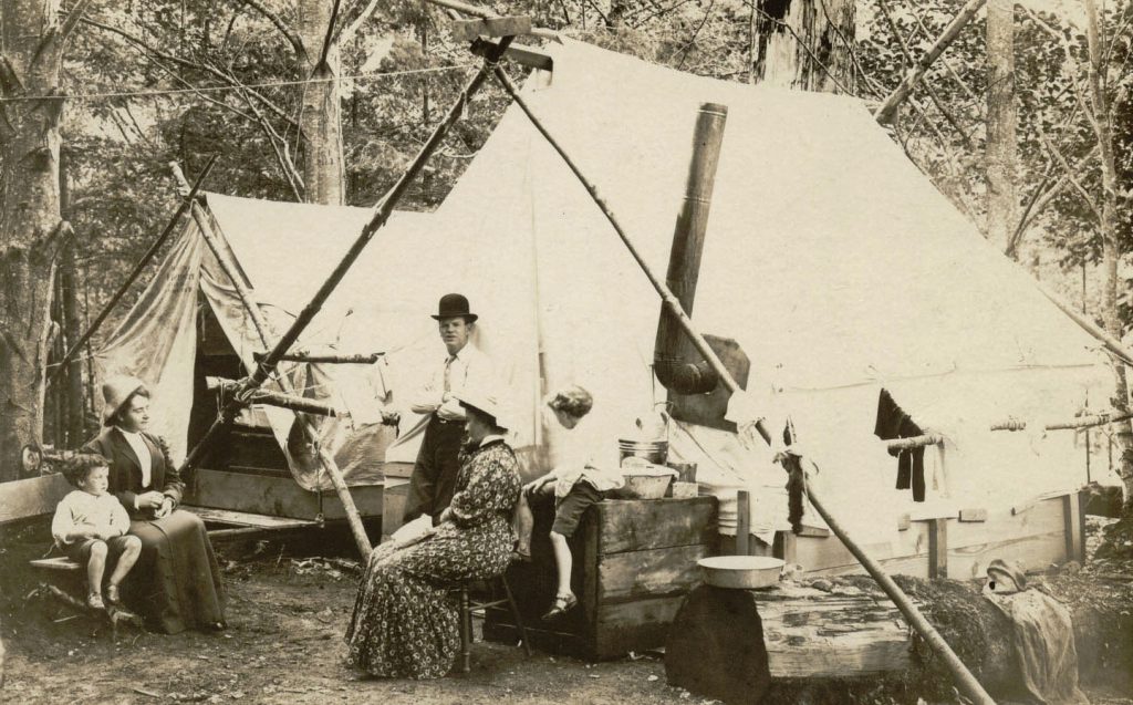 An a-frame canvas tent, an outdoor wooden stove, a man in a bwler hat and woman in early 20th century dress in the woods on Bowen Island BC
