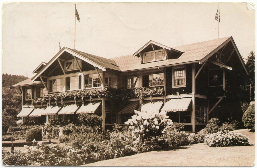 A stately wooden hotel with striped canvas awnings and a flower garden in front.