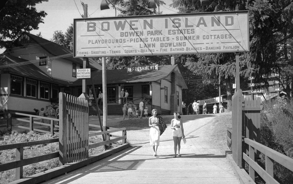 two young women walk on the wide wooden bridge under a sign which reads Bowen Island - Bowen Park Estates - Playgrounds, picnic tables, lawn bowling, summer cottages